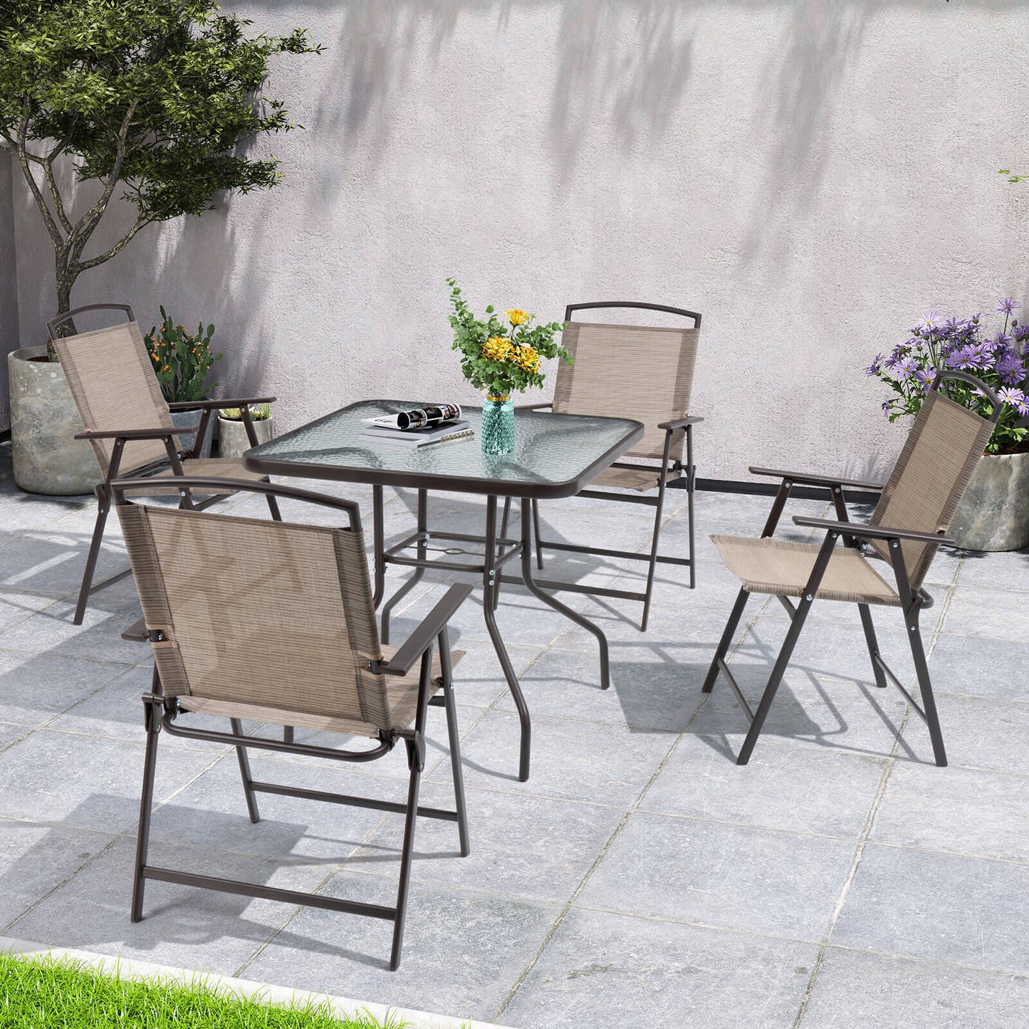5-piece outdoor patio dining set with glass table and folding chairs setup on a stone paved backyard.