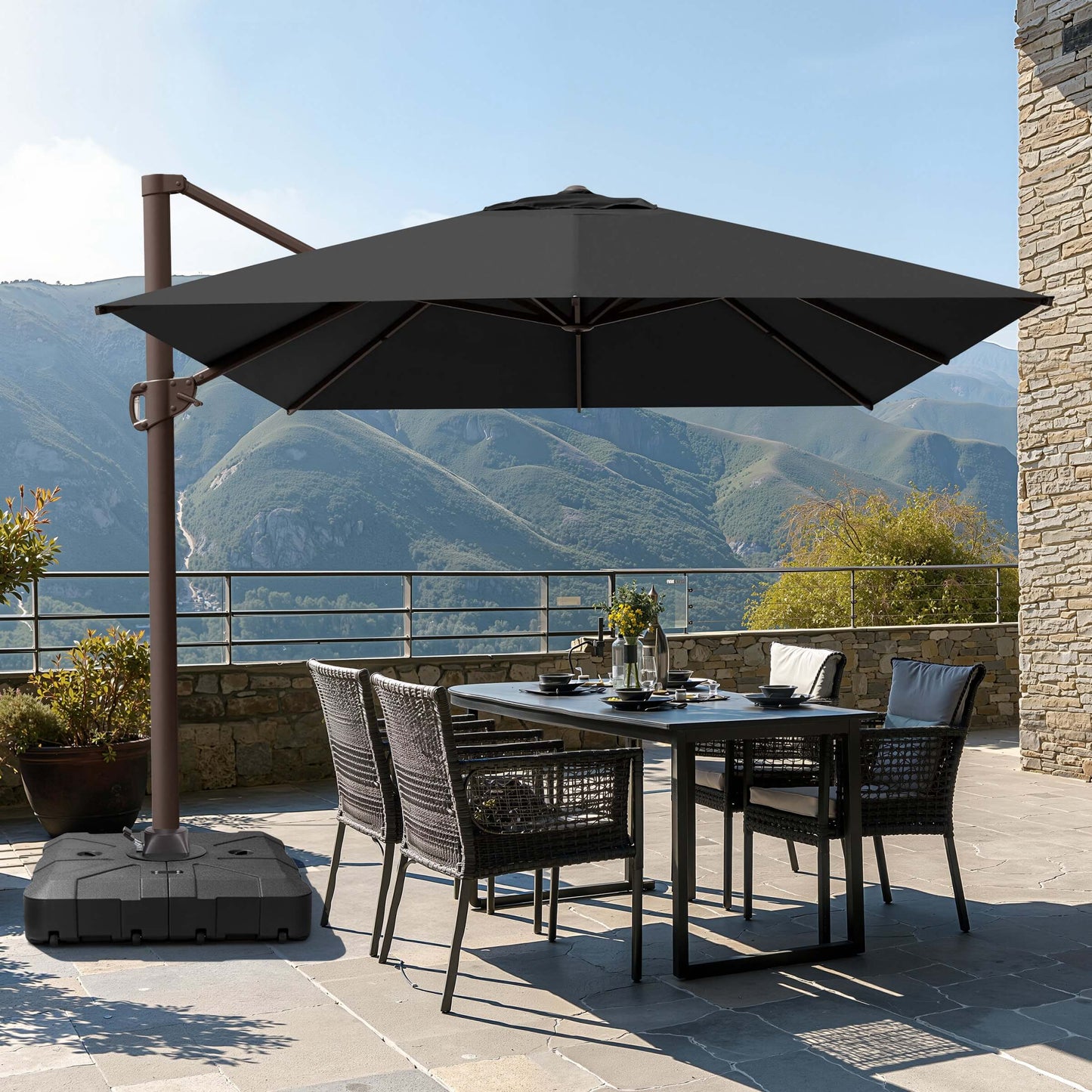 Dark gray square cantilever patio umbrella with wheeled base shading a wicker dining set on a scenic mountain terrace.