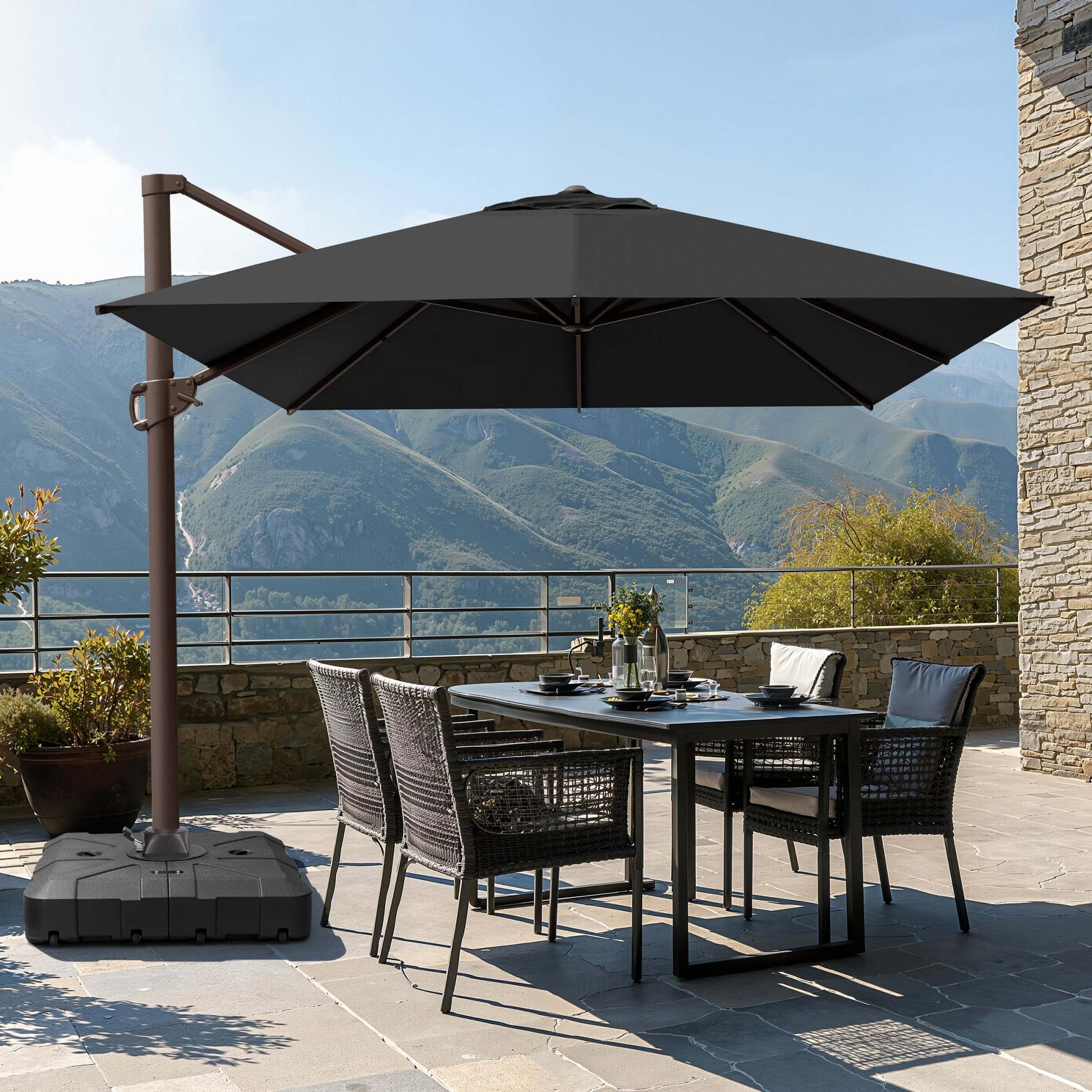 Dark gray square cantilever patio umbrella with wheeled base shading a wicker dining set on a scenic mountain terrace.