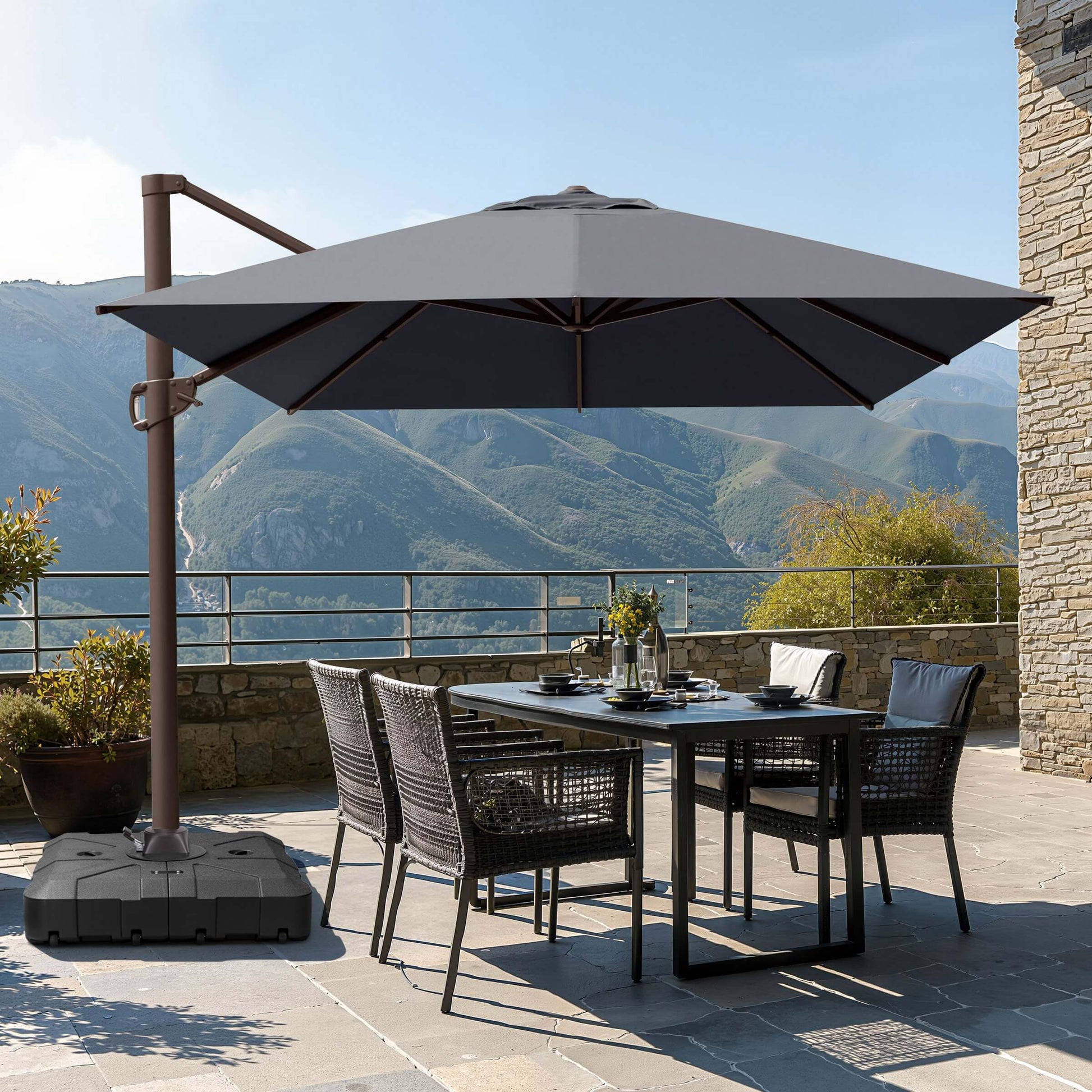 Gray square cantilever patio umbrella with wheeled base shading a wicker dining set on a scenic mountain terrace.