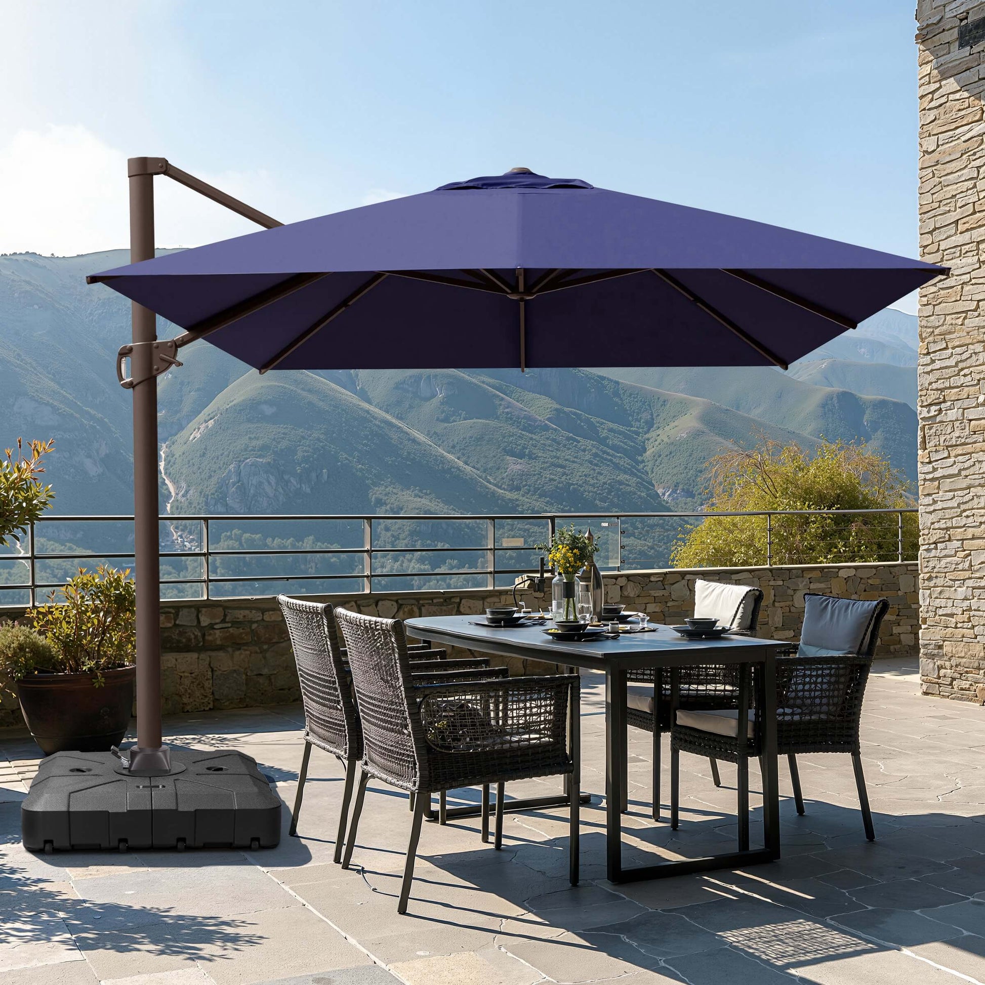 Navy blue square cantilever patio umbrella with wheeled base shading a wicker dining set on a mountain terrace.