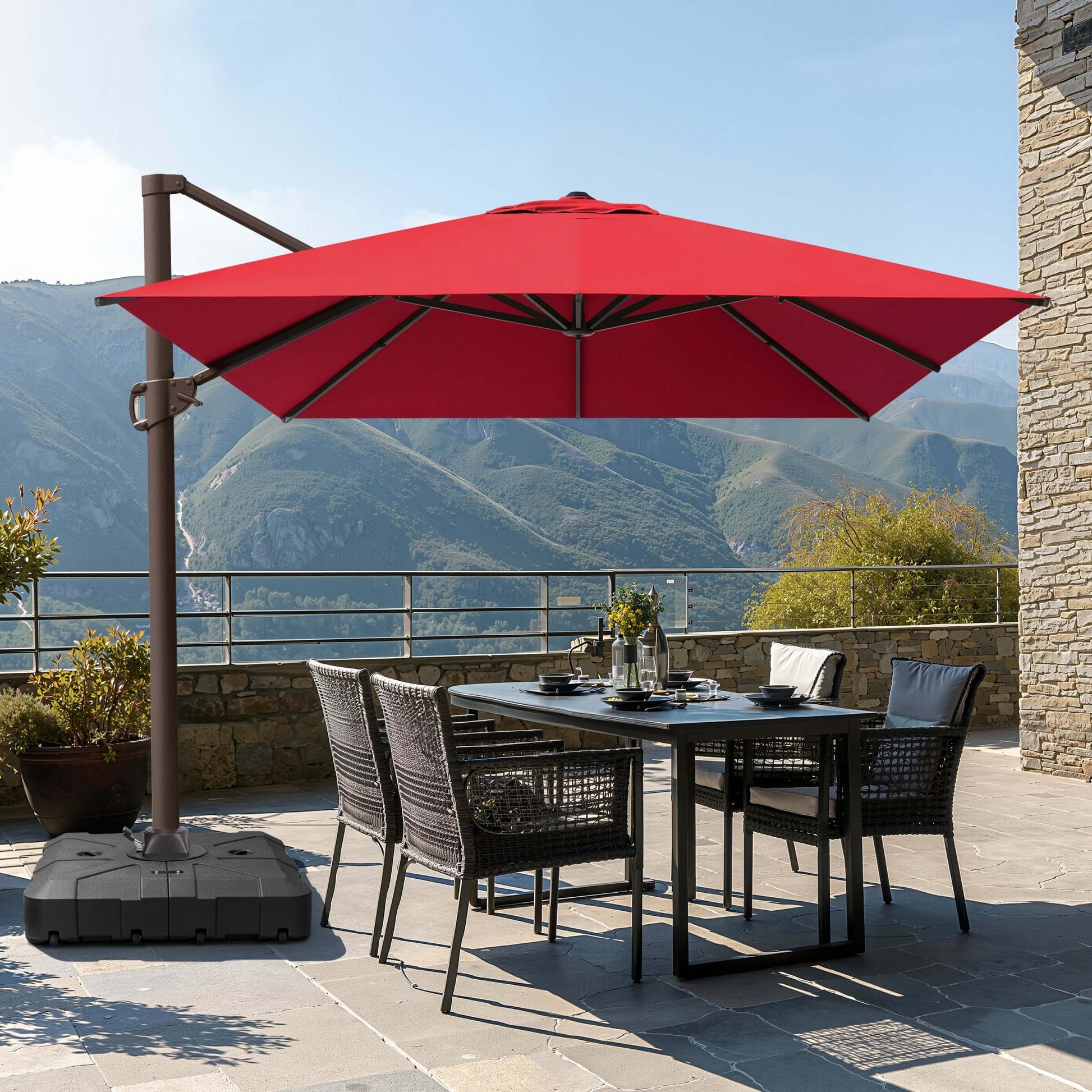 Red square cantilever patio umbrella with wheeled base shading a wicker dining set on a mountain terrace.