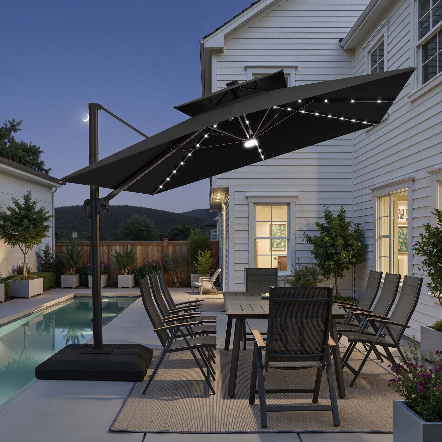Tilted 12x9 ft black lighted cantilever umbrella with low-profile base shading a poolside dining set at twilight