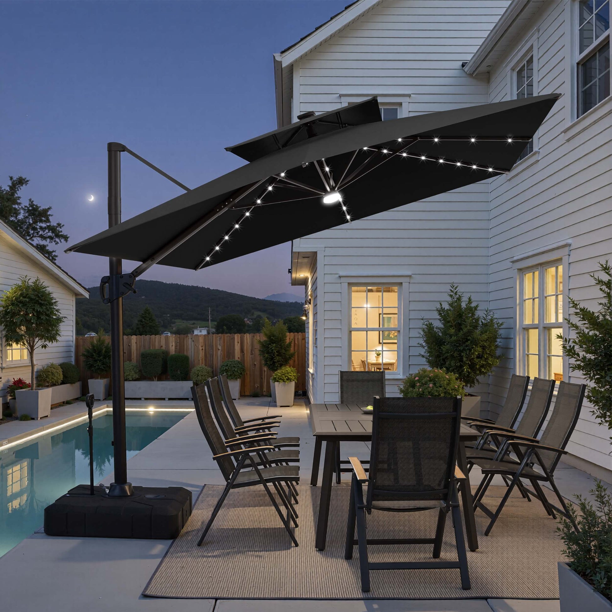 Tilted 12x9 ft black lighted cantilever umbrella with wheeled base shading a poolside dining set at twilight