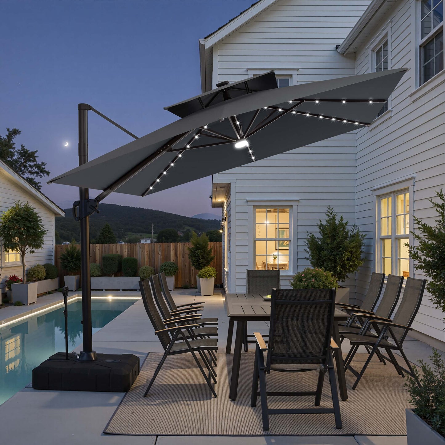 Tilted 12x9 ft grey lighted cantilever umbrella with wheeled base shading a poolside dining set at twilight