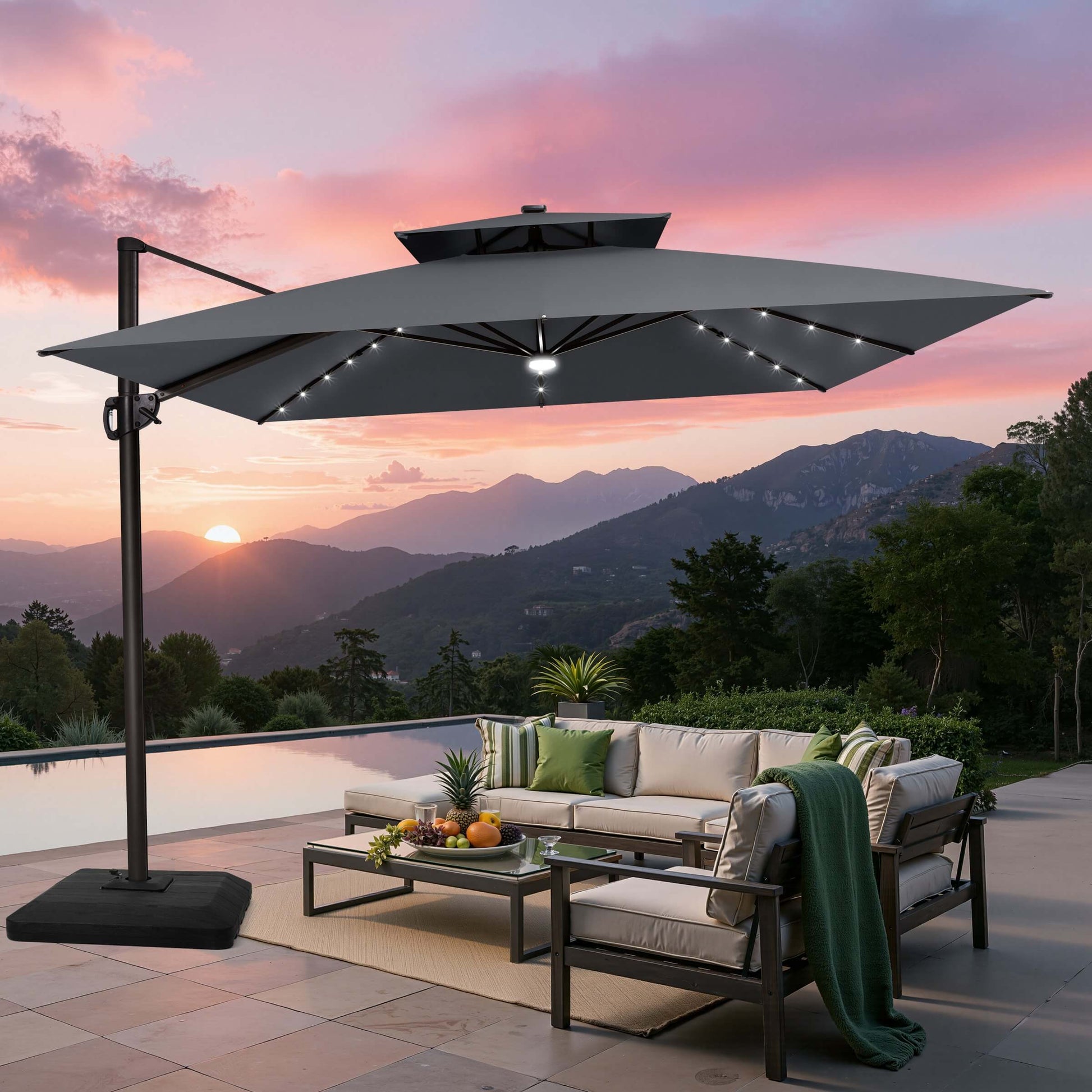 Grey double top LED cantilever umbrella with base over patio furniture set by poolside with mountain view sunset.