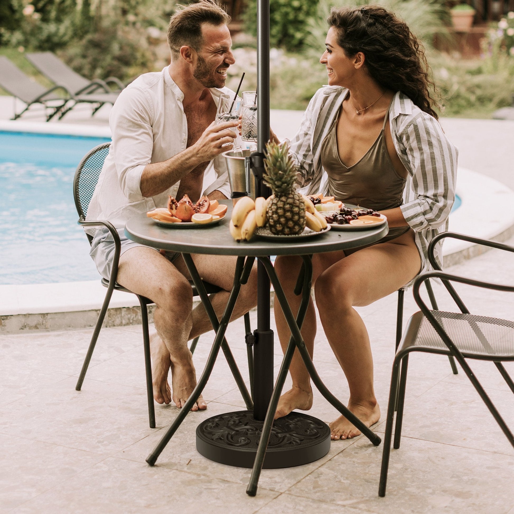 Black 30-46 lbs round floral cement umbrella base under a patio table with a happy couple by the pool.